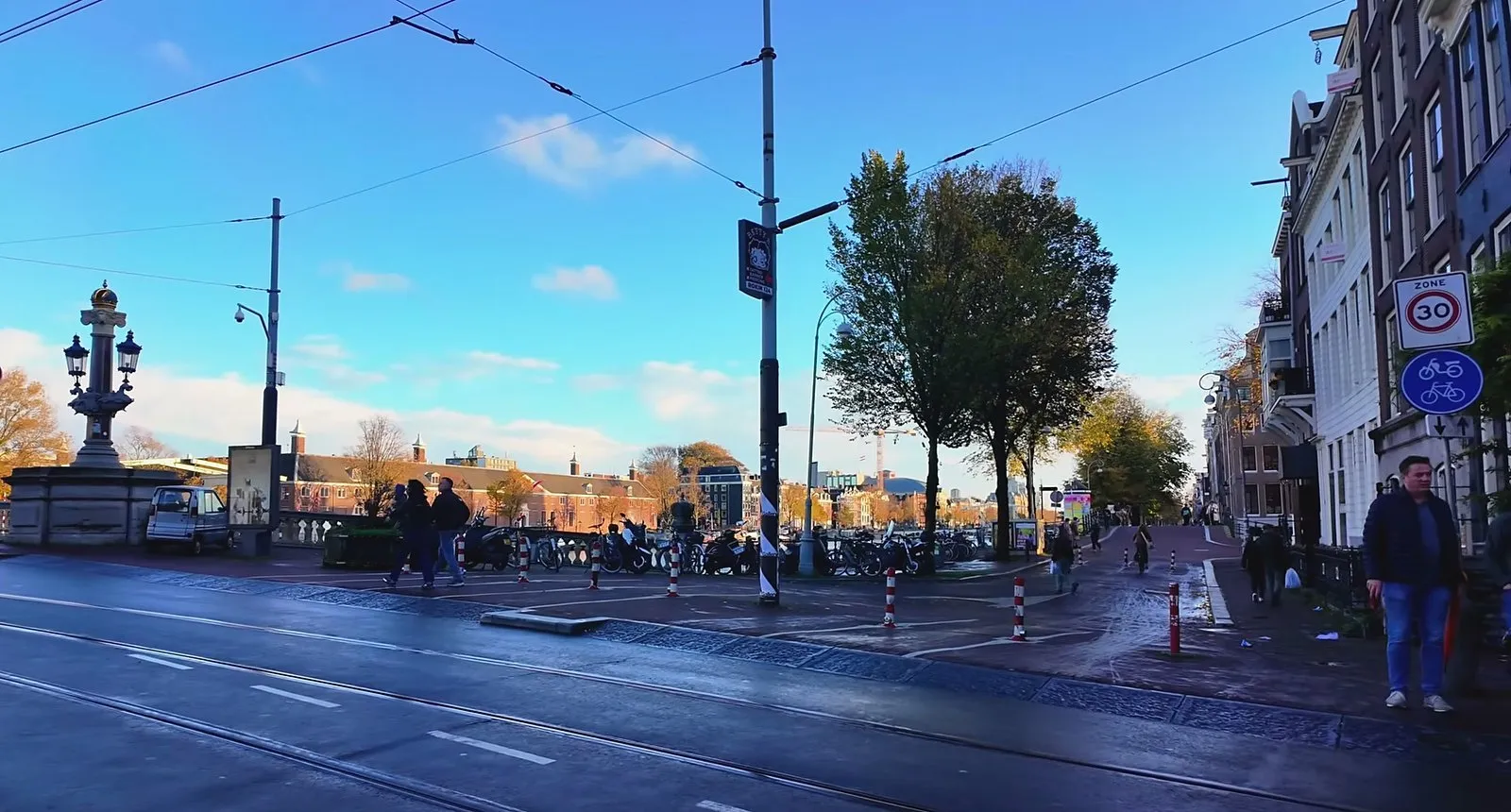 Golden late-afternoon light on a bridge and tree-lined canal near the Amstel in Amsterdam, under a blue sky with scattered clouds