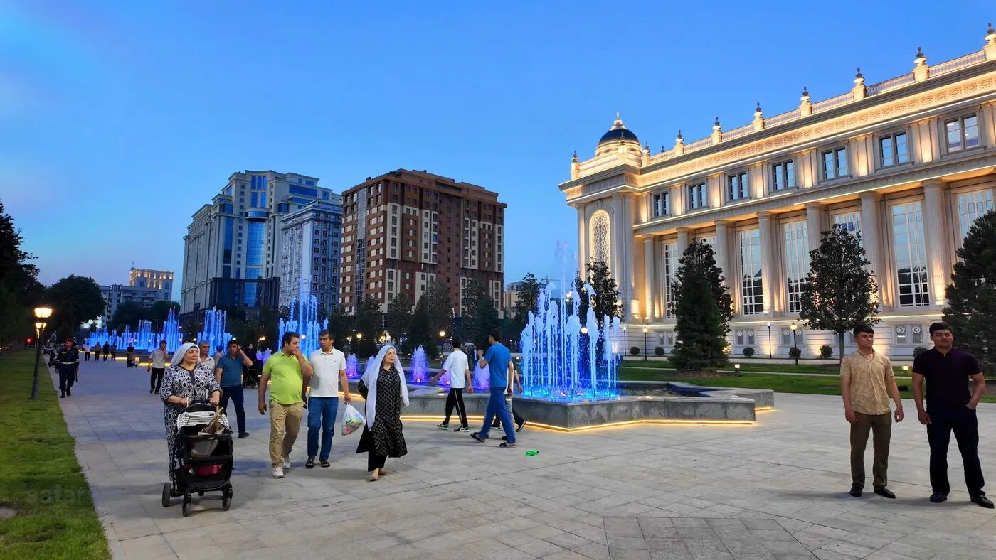 Families walking past lit fountains and a palace facade at blue hour in Dushanbe
