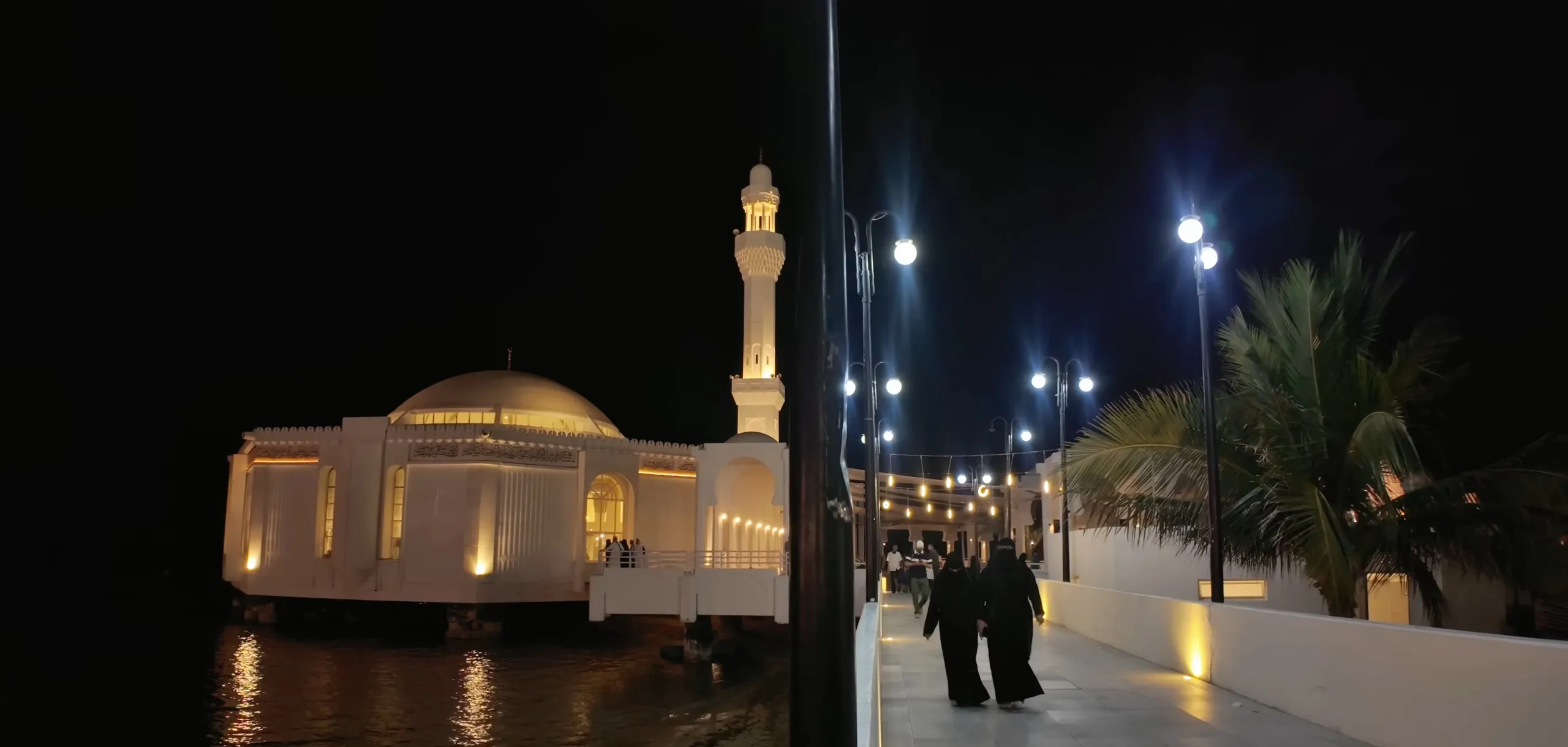 Al-Rahmah Mosque on its pillars over the Red Sea at night, its lights reflected in the water, beside the lamp-lined walkway