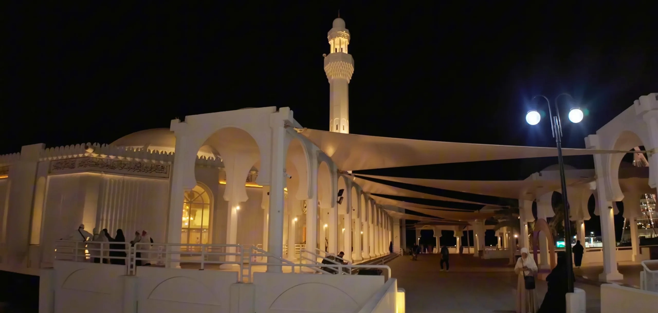 White arches of the arcade around Al-Rahmah Mosque in Jeddah, lit warm at night under shade sails, with the minaret above