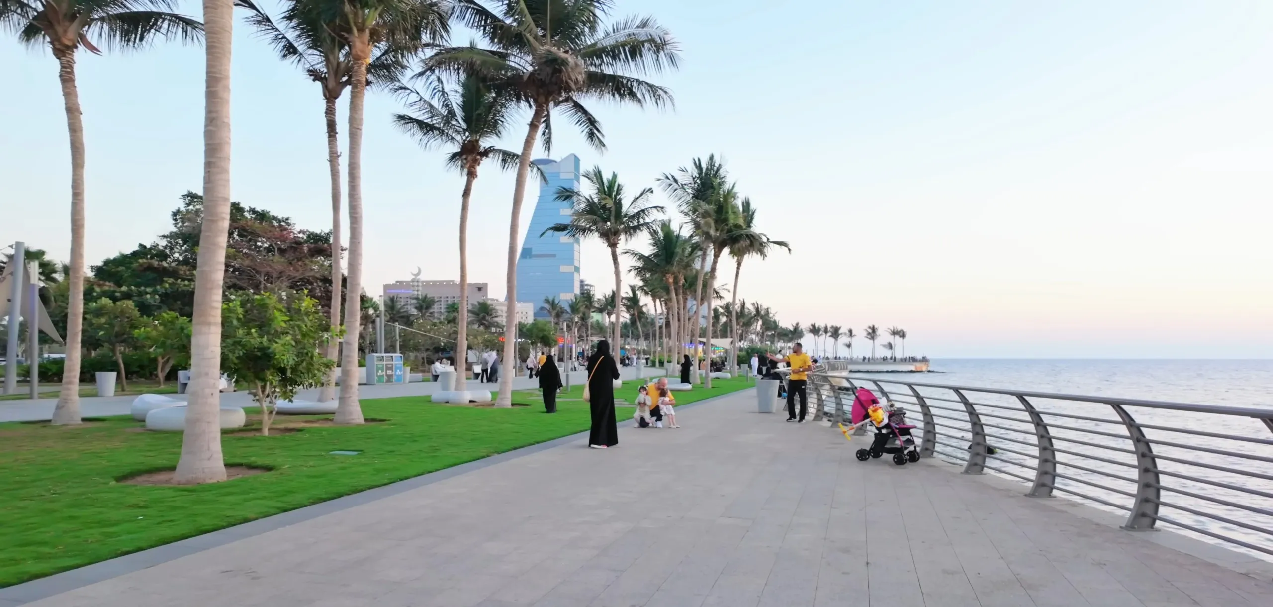 The palm-lined promenade of the Jeddah Corniche just after sunset, with people walking by the sea railing, a small group at the edge of the lawn and a tall blue glass tower behind