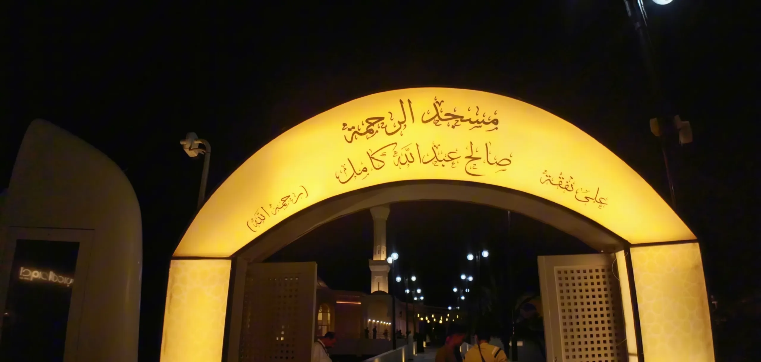 The lit entrance arch on the walkway to Al-Rahmah Mosque in Jeddah, its name written in Arabic across the top, with the minaret seen through the arch