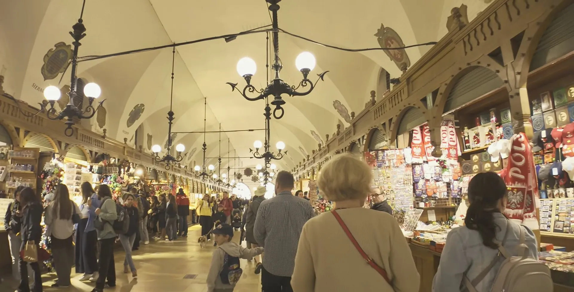 The long vaulted hall inside Kraków’s Cloth Hall, lined with wooden market stalls selling souvenirs and amber under hanging lamps and painted coats of arms