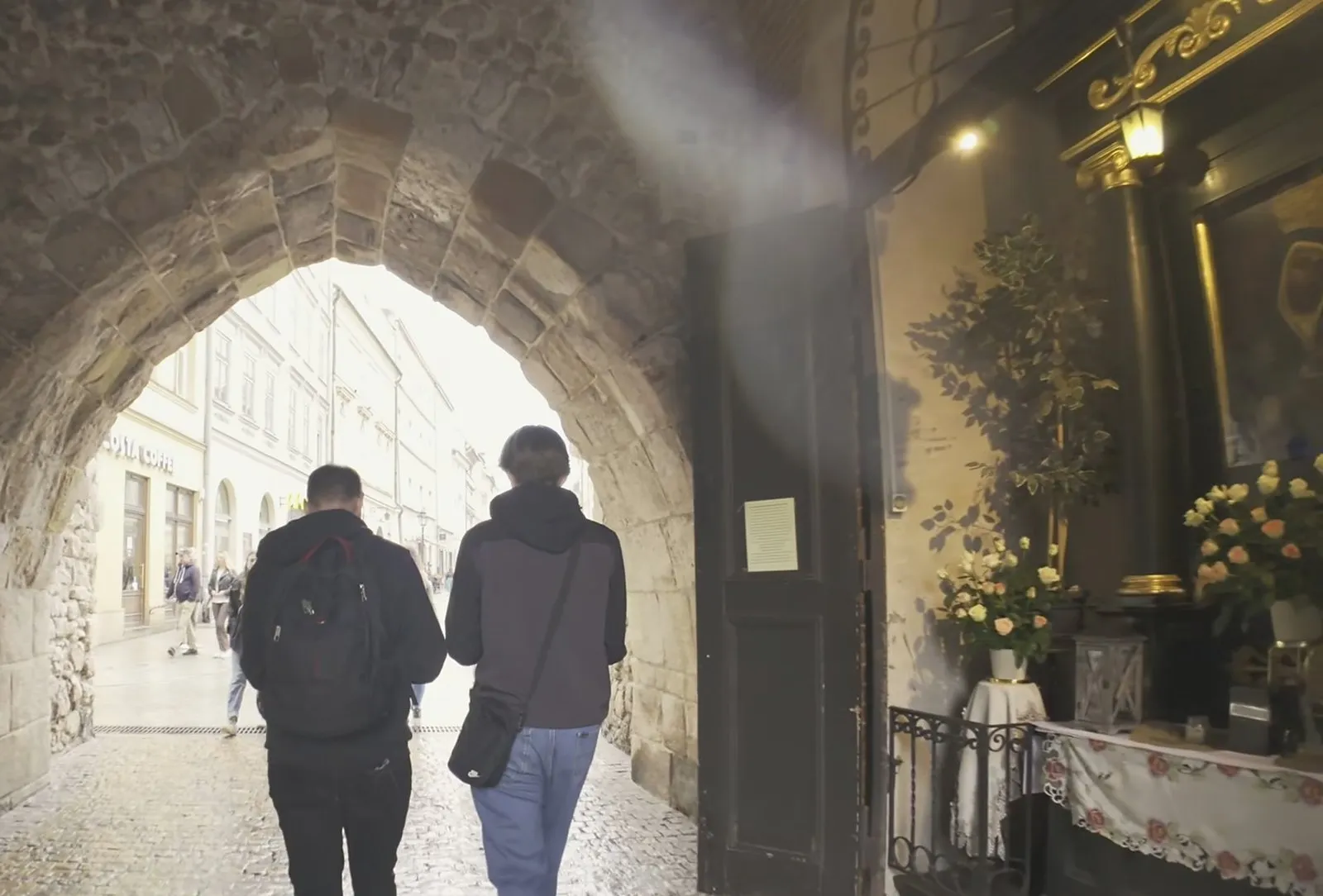 Walking through the stone archway of St Florian’s Gate toward Floriańska Street, with a lit shrine and fresh flowers set into the wall on the right