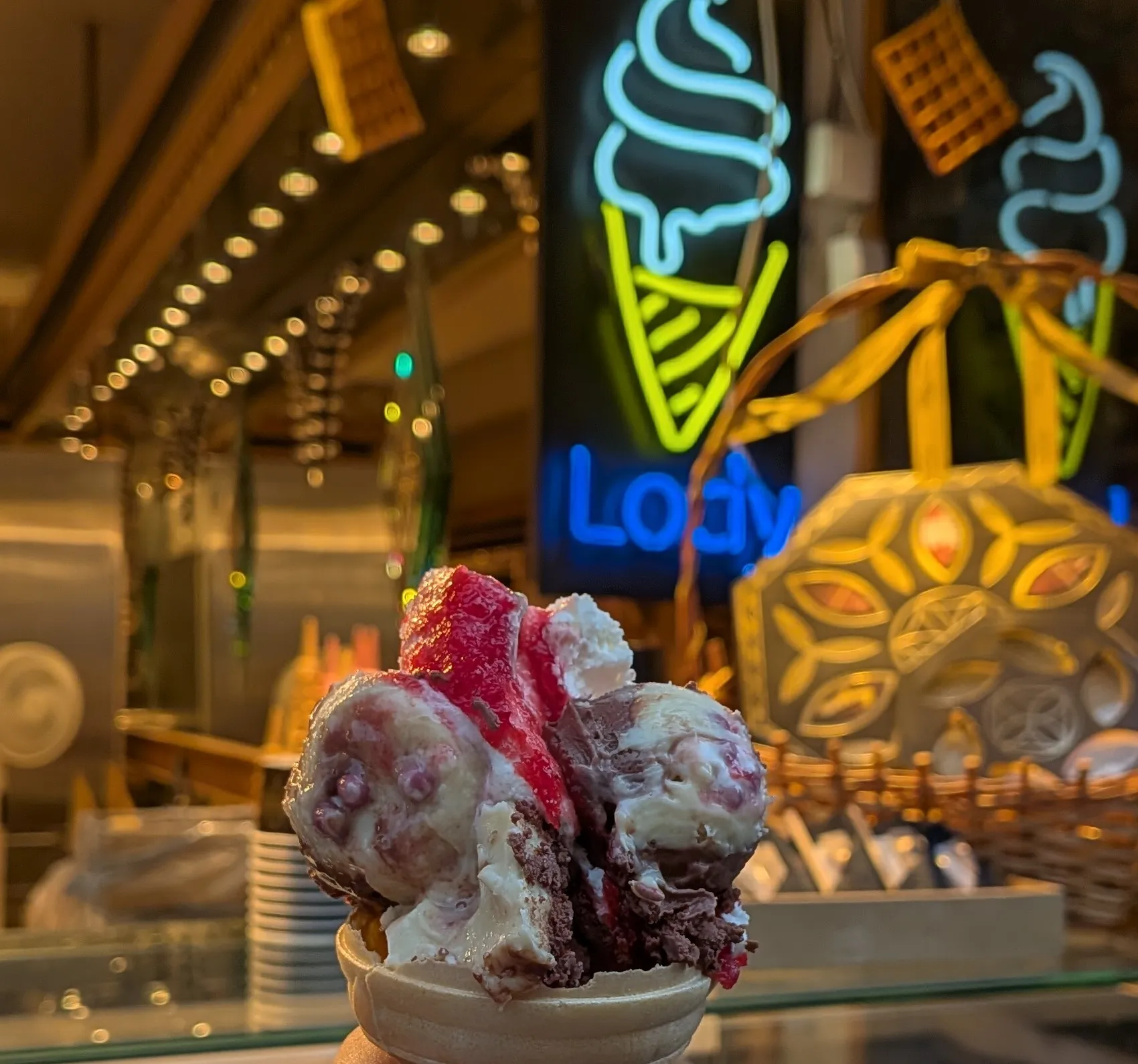 A cone of pink and chocolate ice cream held up in front of a glowing neon ice-cream sign
