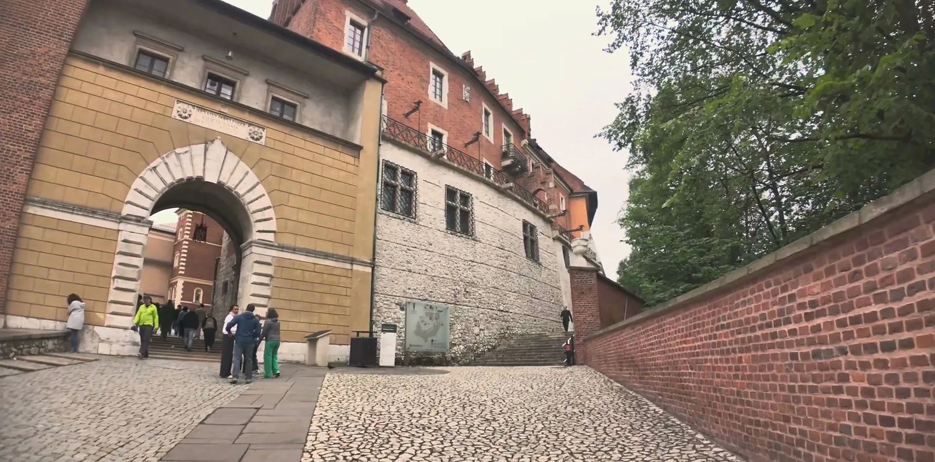 The cobbled ramp up to Wawel Castle, with a red-brick wall on the right and an arched stone gateway ahead leading into the castle grounds