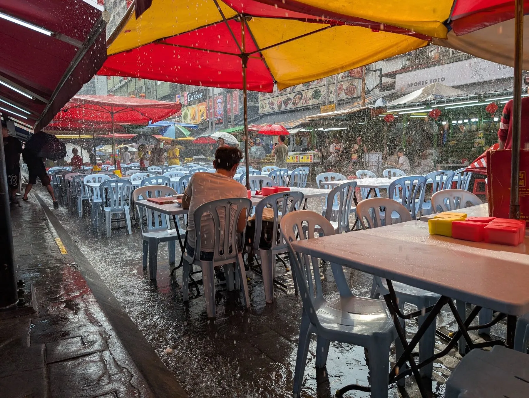 Heavy rain falling on red and yellow umbrellas over a street-food lane in Kuala Lumpur