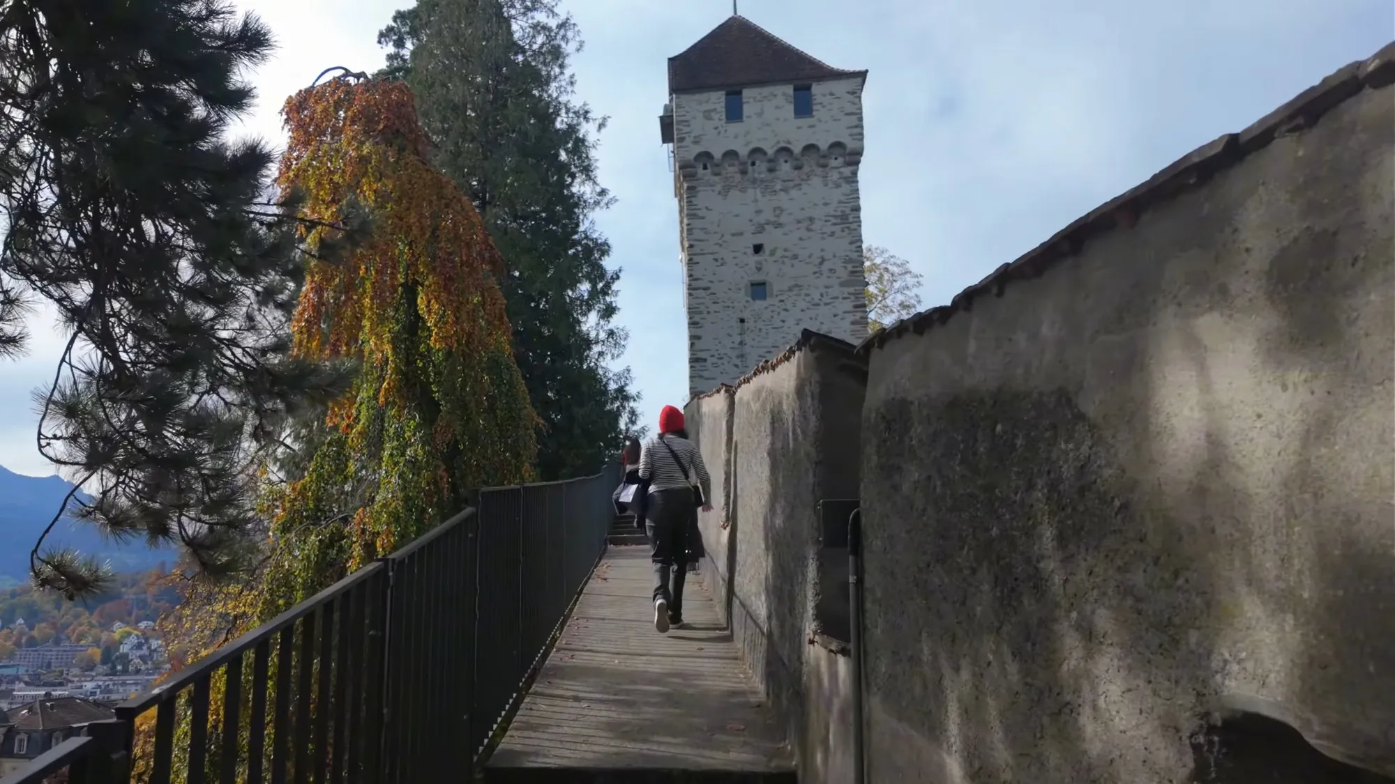 A stone tower with a pointed roof on the Musegg Wall in Lucerne, above the wall walk and a tree in autumn colours, under a blue sky