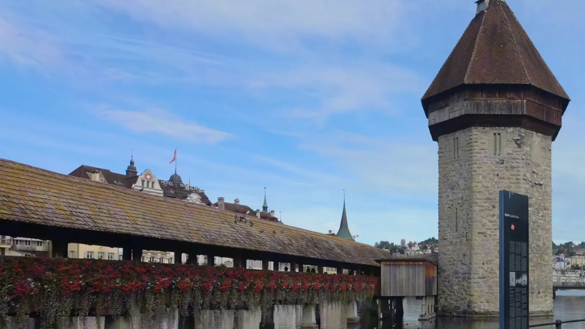 The Chapel Bridge in Lucerne with red flowers along its side and the octagonal stone Water Tower beside it in the Reuss, under a blue sky