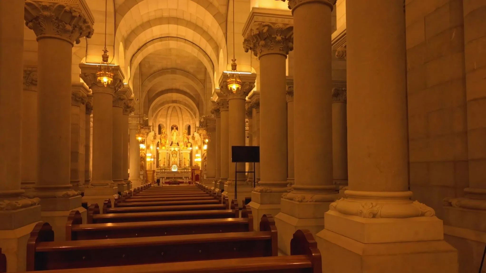 The crypt of the Almudena Cathedral in Madrid, with rows of thick stone columns, wooden pews and a gold-lit altar at the end of the nave