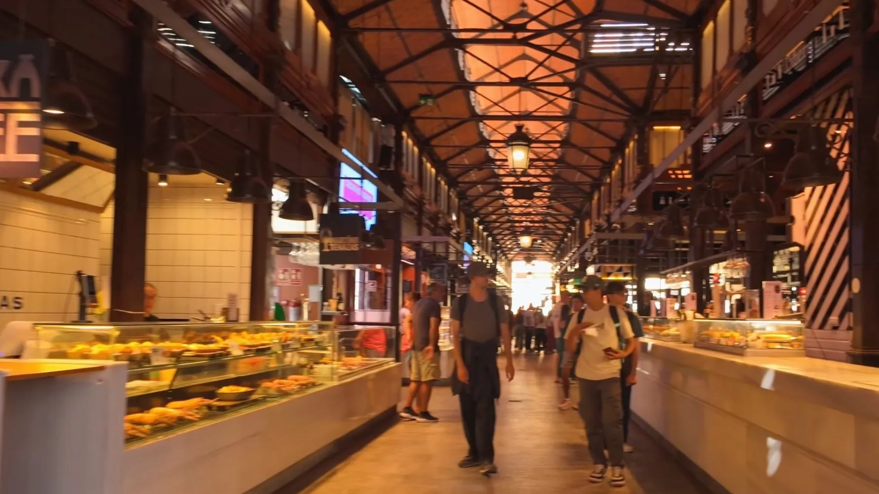The long hall of the Mercado de San Miguel in Madrid, with glass food counters on both sides and an iron roof lit warm orange overhead
