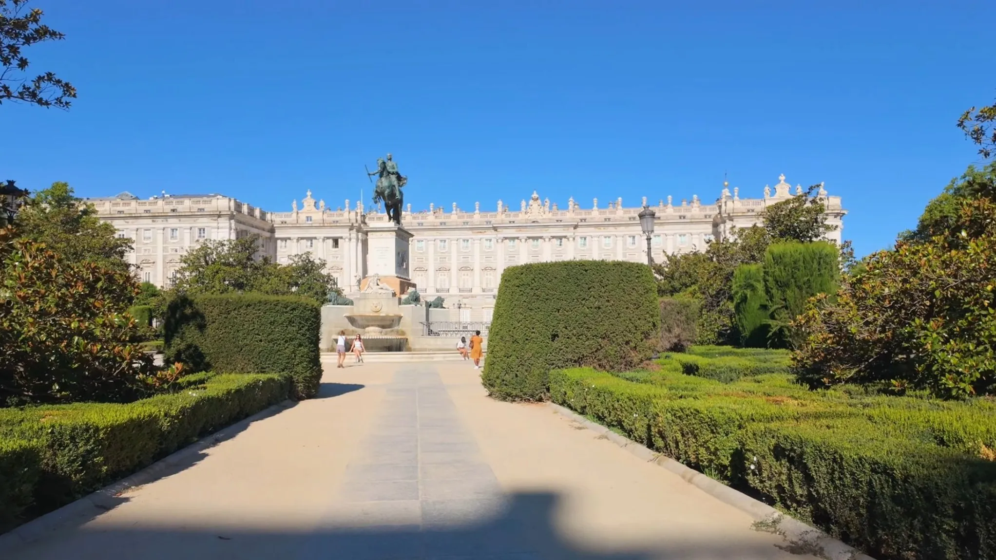 The Royal Palace of Madrid at the end of a garden path in Plaza de Oriente, with the bronze equestrian statue of Philip IV in front of it and clipped hedges on both sides