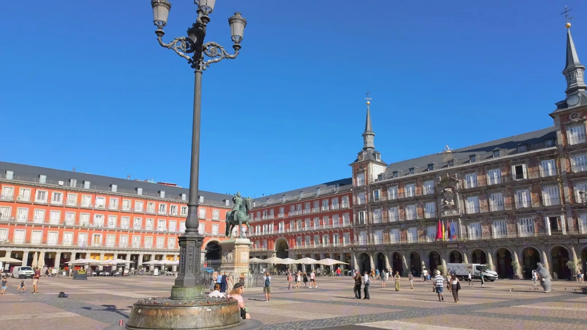 Plaza Mayor in Madrid on a sunny morning, with red arcaded buildings, the painted Casa de la Panadería with its two spires, and the bronze statue of Philip III in the center