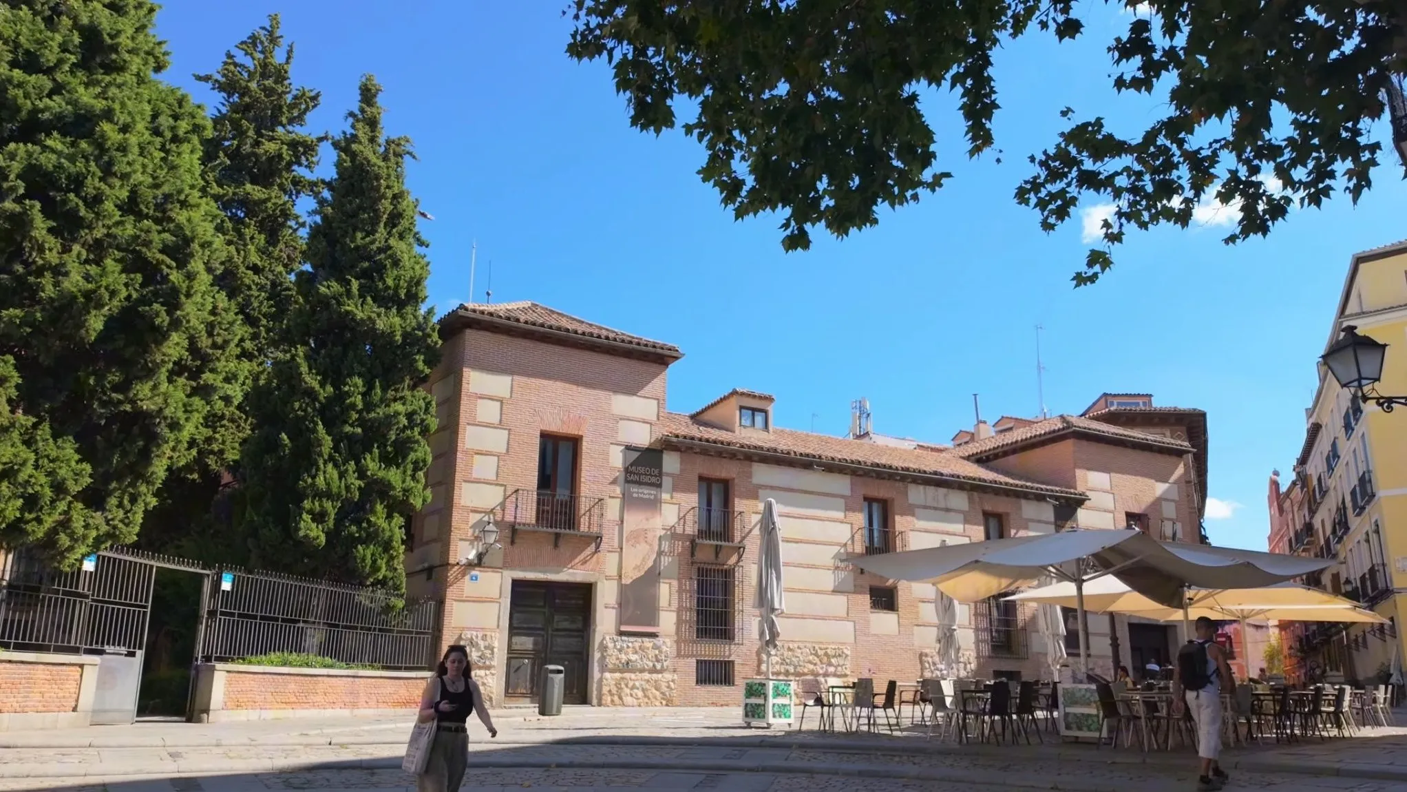 Plaza de San Andrés in La Latina, Madrid, with the brick and stone Museo de San Isidro behind a terrace of café umbrellas and tall dark trees on the left