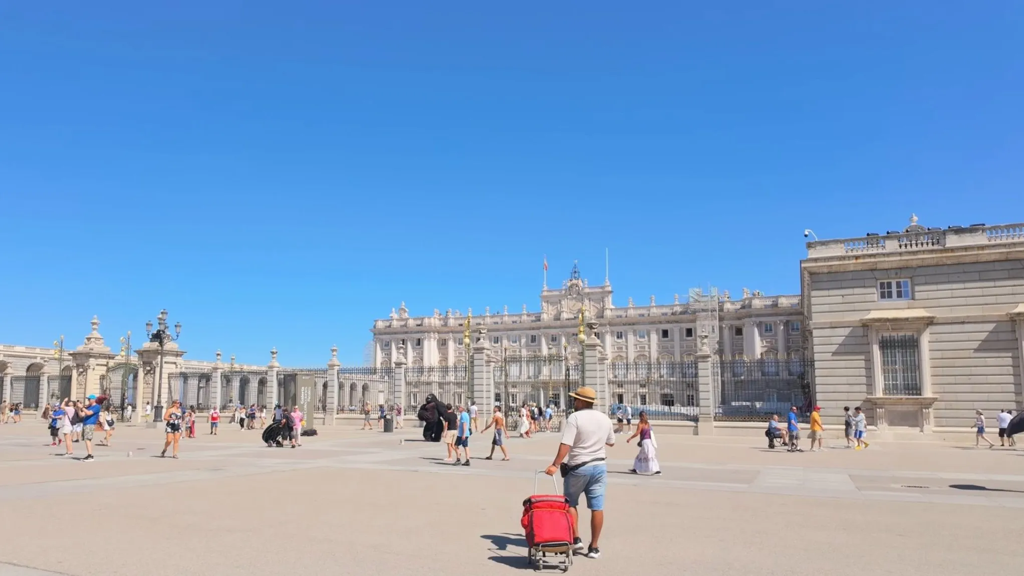 The Royal Palace of Madrid seen across the open Plaza de la Armería, with visitors crossing the pale stone square under a clear blue sky
