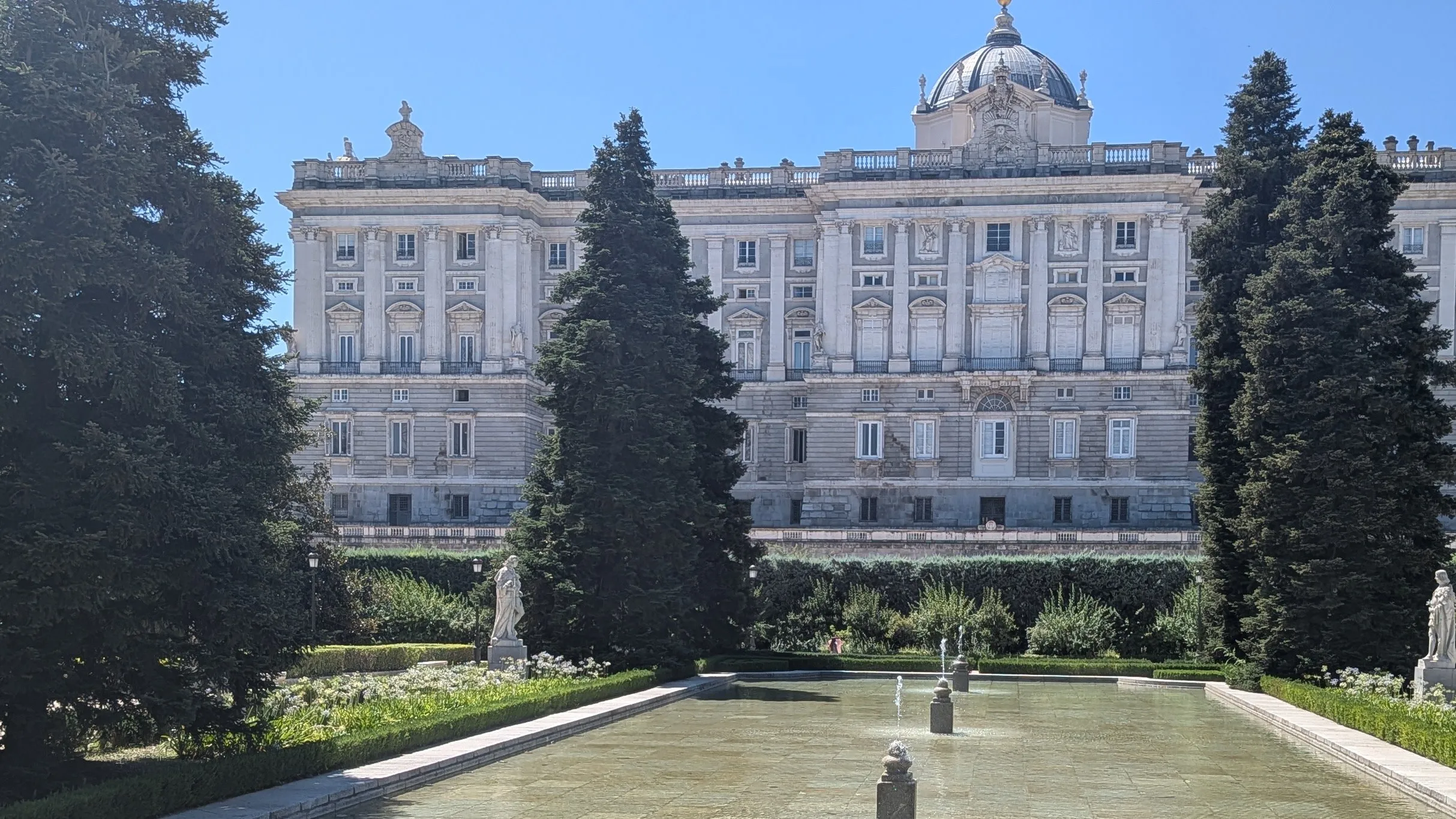 The north front of the Royal Palace of Madrid behind the formal pool, hedges and tall dark trees of the Sabatini Gardens