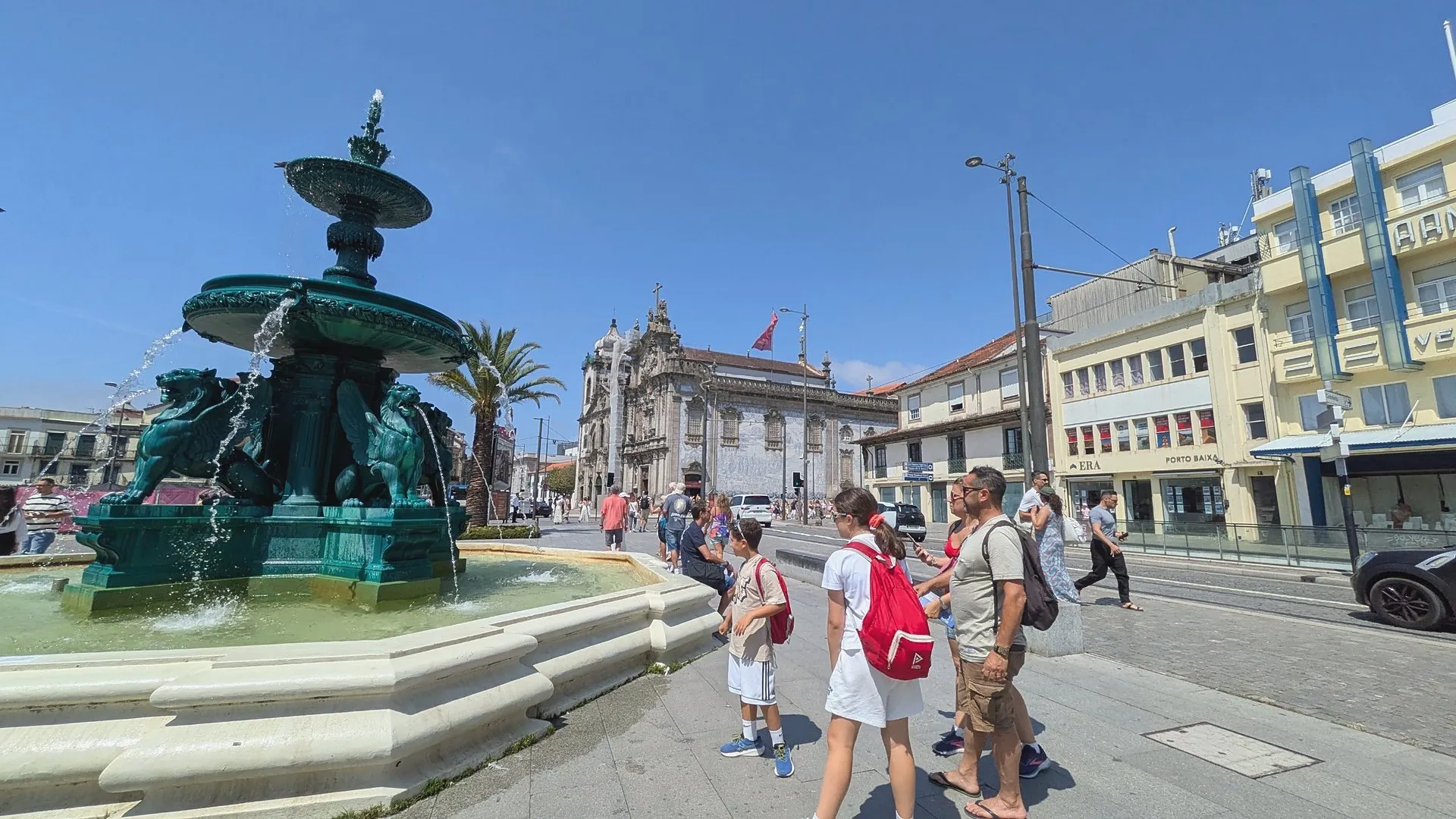 The dark green Lions Fountain spraying water in Praça de Gomes Teixeira, with a family looking at a map in front of it and the azulejo-tiled side of Igreja do Carmo behind