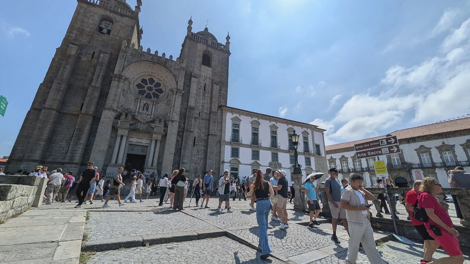 A busy Terreiro da Sé square in full sun, with crowds walking past the granite facade and rose window of Porto Cathedral and a brown sign pointing to Igreja dos Grilos and Arte Sacra