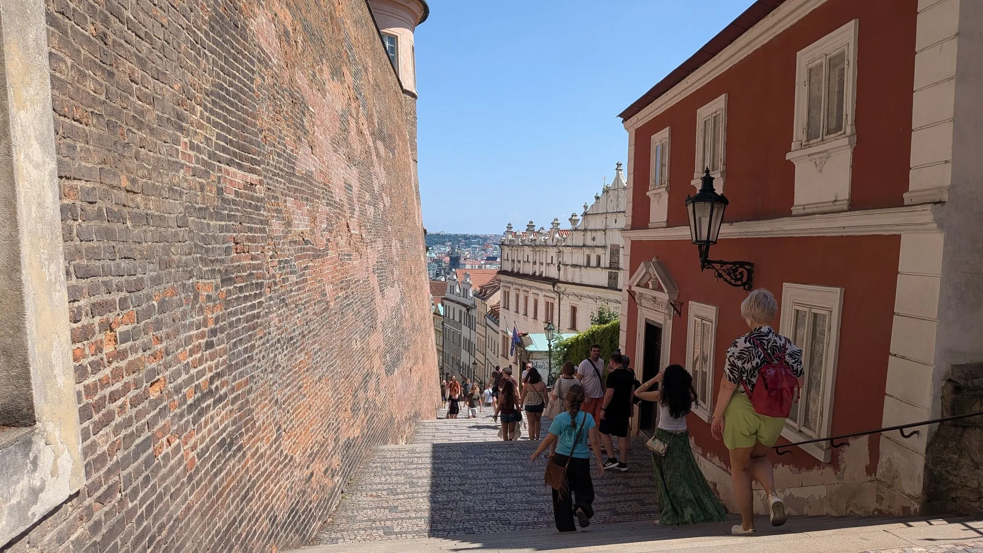 A long stepped lane leading down from Prague Castle between an old brick wall and a red house with a lantern, with people walking down ahead and the rooftops of the city below