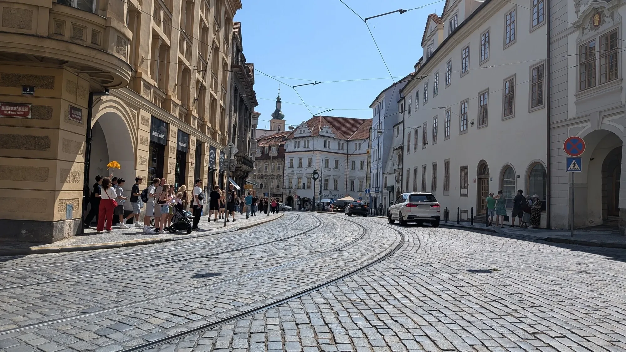 Sunlit cobbles and curving tram lines on Malostranské náměstí in Prague’s Lesser Town, between an ochre corner building with arcades and a row of pale old house fronts, with a church tower in the distance
