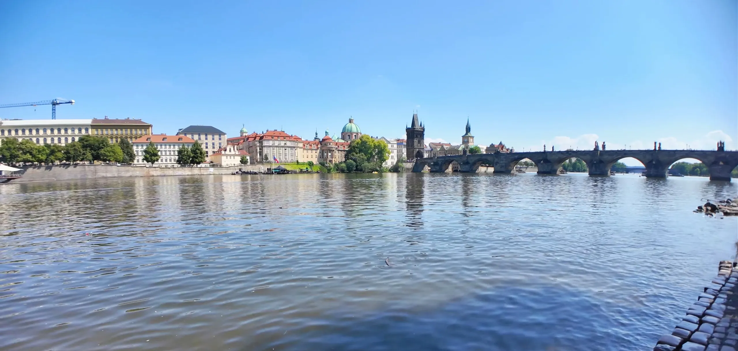 Charles Bridge and the Old Town Bridge Tower across the Vltava in Prague on a clear summer afternoon, seen from the Lesser Town riverbank, with the domes and towers of the Old Town along the far bank