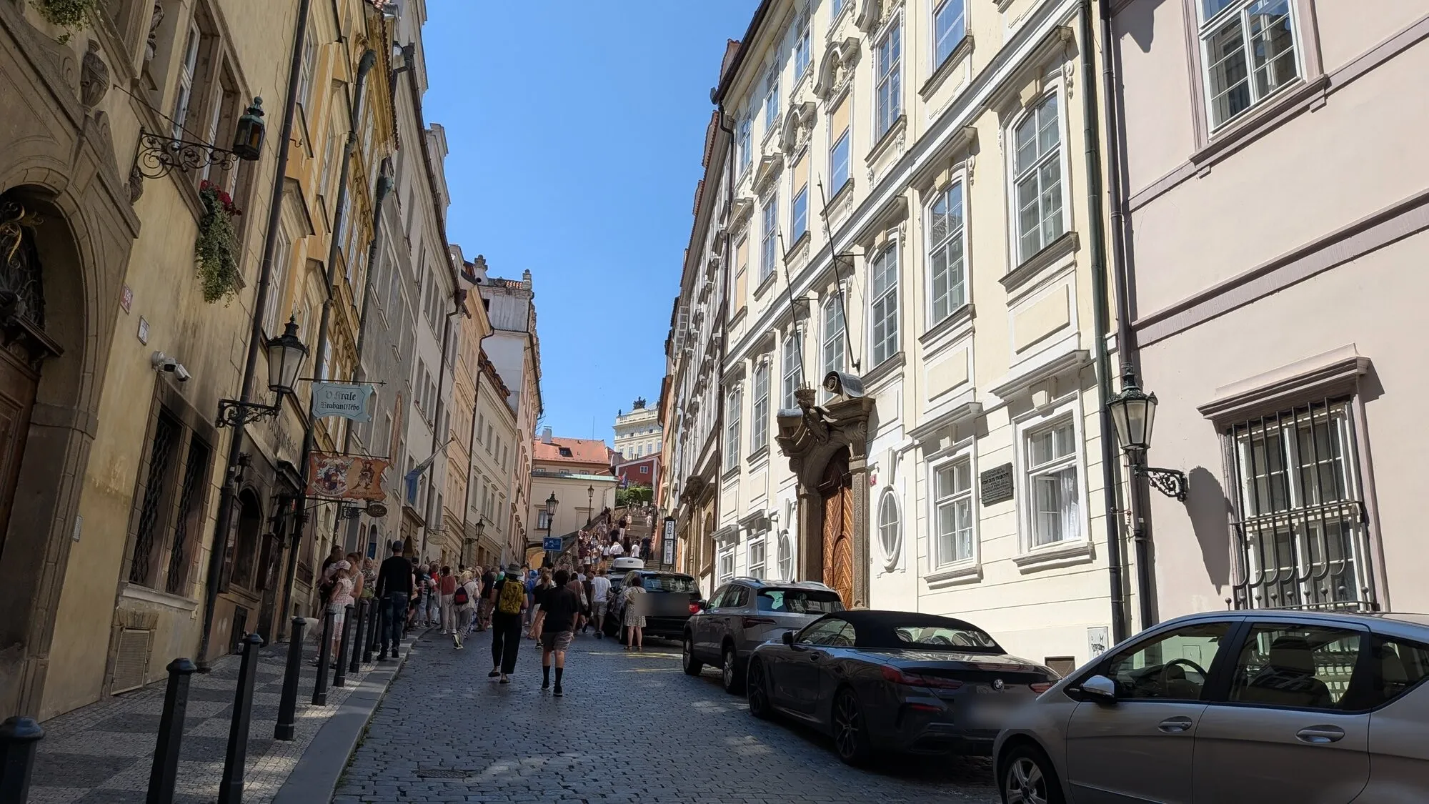 Thunovská in Prague’s Lesser Town on a bright summer day, a cobbled street of pale baroque house fronts with lanterns and hanging signs rising to the crowded foot of the Castle Steps, with people walking up it and parked cars on the right