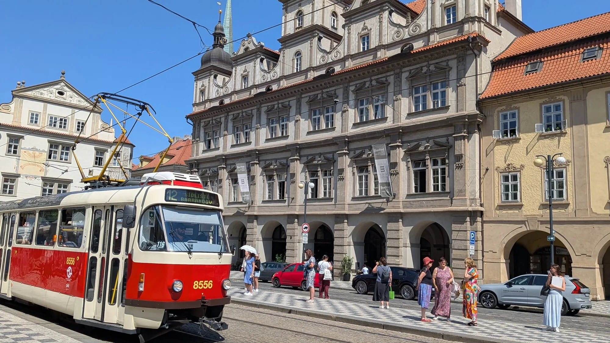A classic red and cream Prague tram on route 12 passing the arcaded Renaissance front of Malostranská beseda on the Lesser Town square, with people waiting at the stop under a clear blue sky