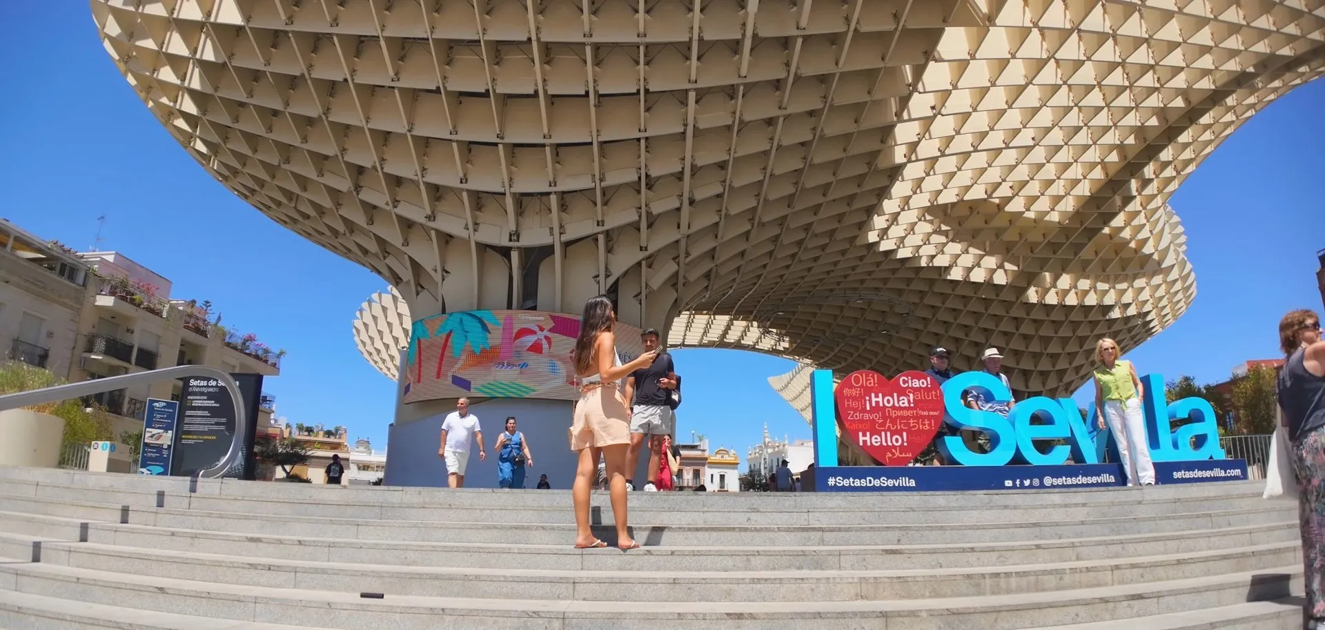 A woman standing on the sunlit steps of Las Setas looking at her phone, with the wooden canopy overhead and people around the I love Sevilla sign
