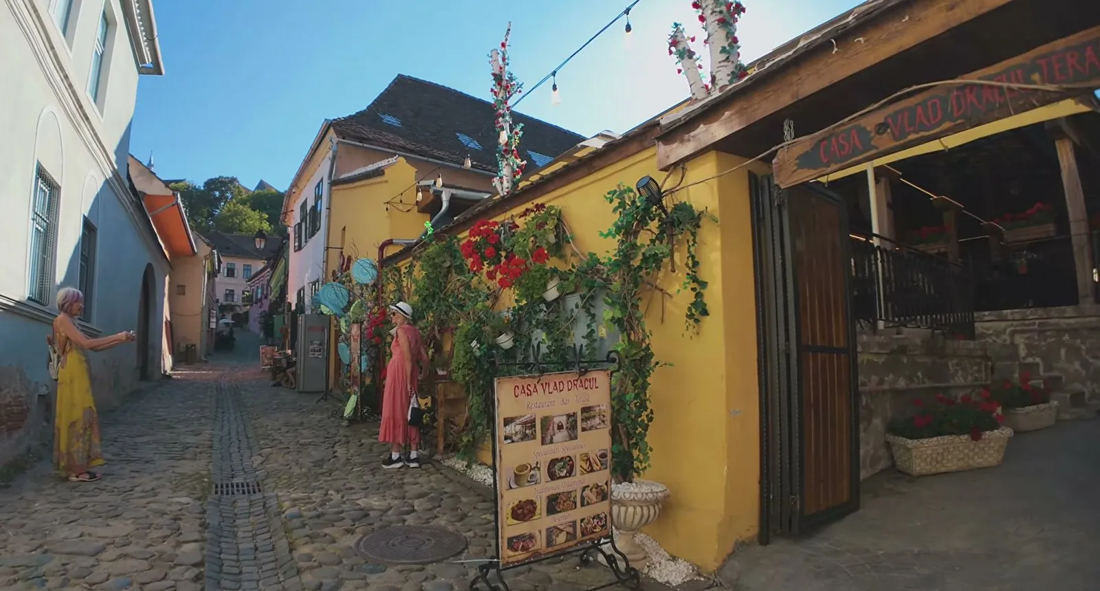 The yellow wall of Casa Vlad Dracul covered in red geraniums and climbing plants, with its wooden sign over the terrace and a cobbled lane running uphill