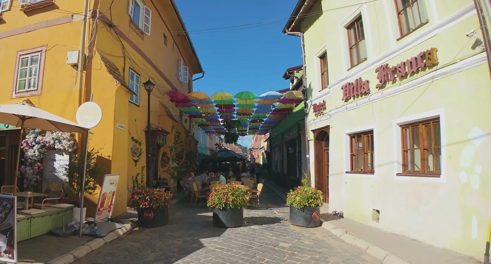 A sunny lower-town lane in Sighișoara with café tables, flower pots and colorful umbrellas hung overhead