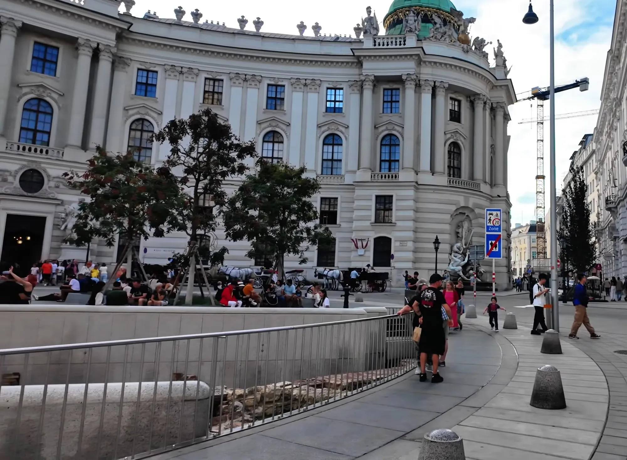 The open Roman excavation behind a metal railing on Michaelerplatz in Vienna, with the green-domed curved front of the Hofburg beyond and pedestrians on the paving