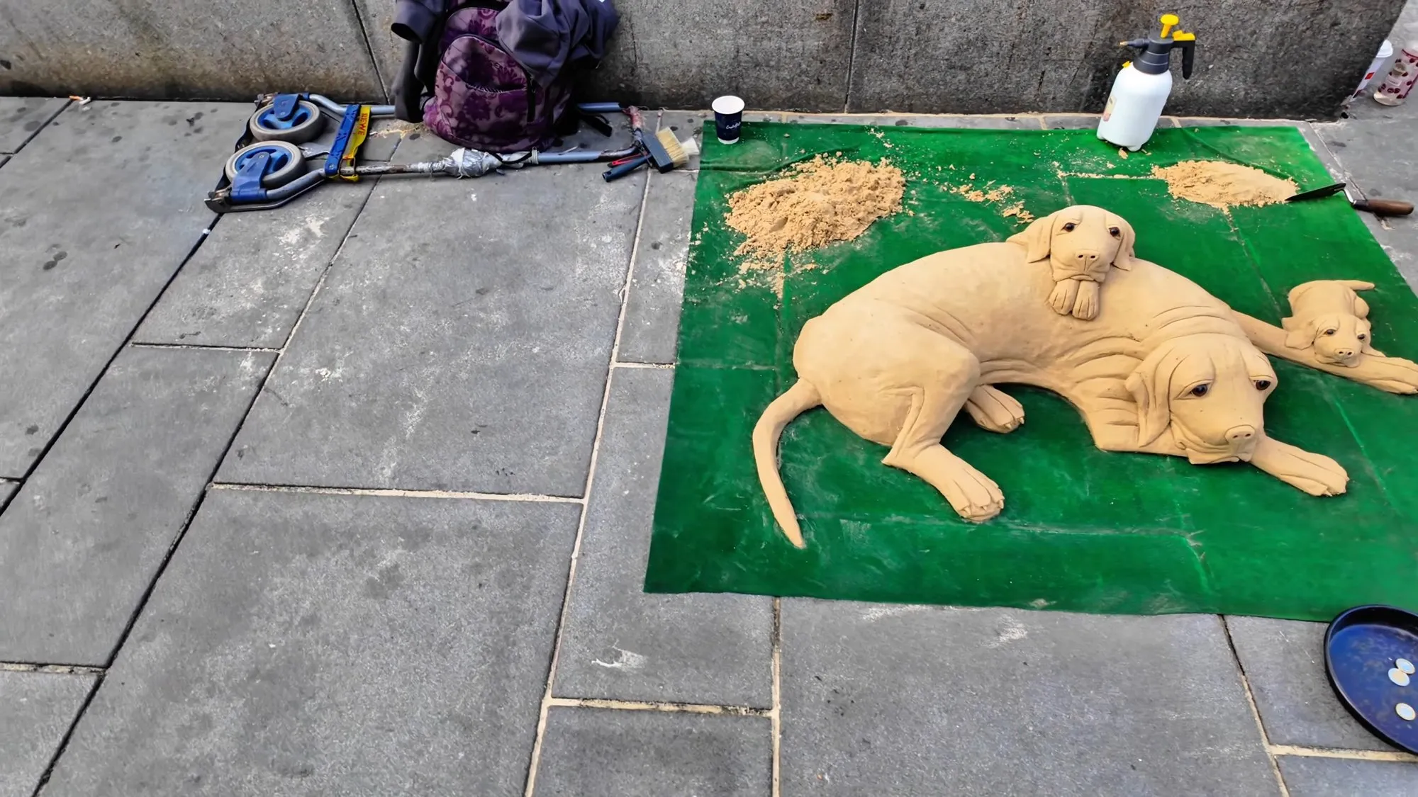 A sand sculpture of a large dog with two puppies, one resting on its back, on a green mat on the paving of the Graben in Vienna, with the sculptor’s tools and bag beside it