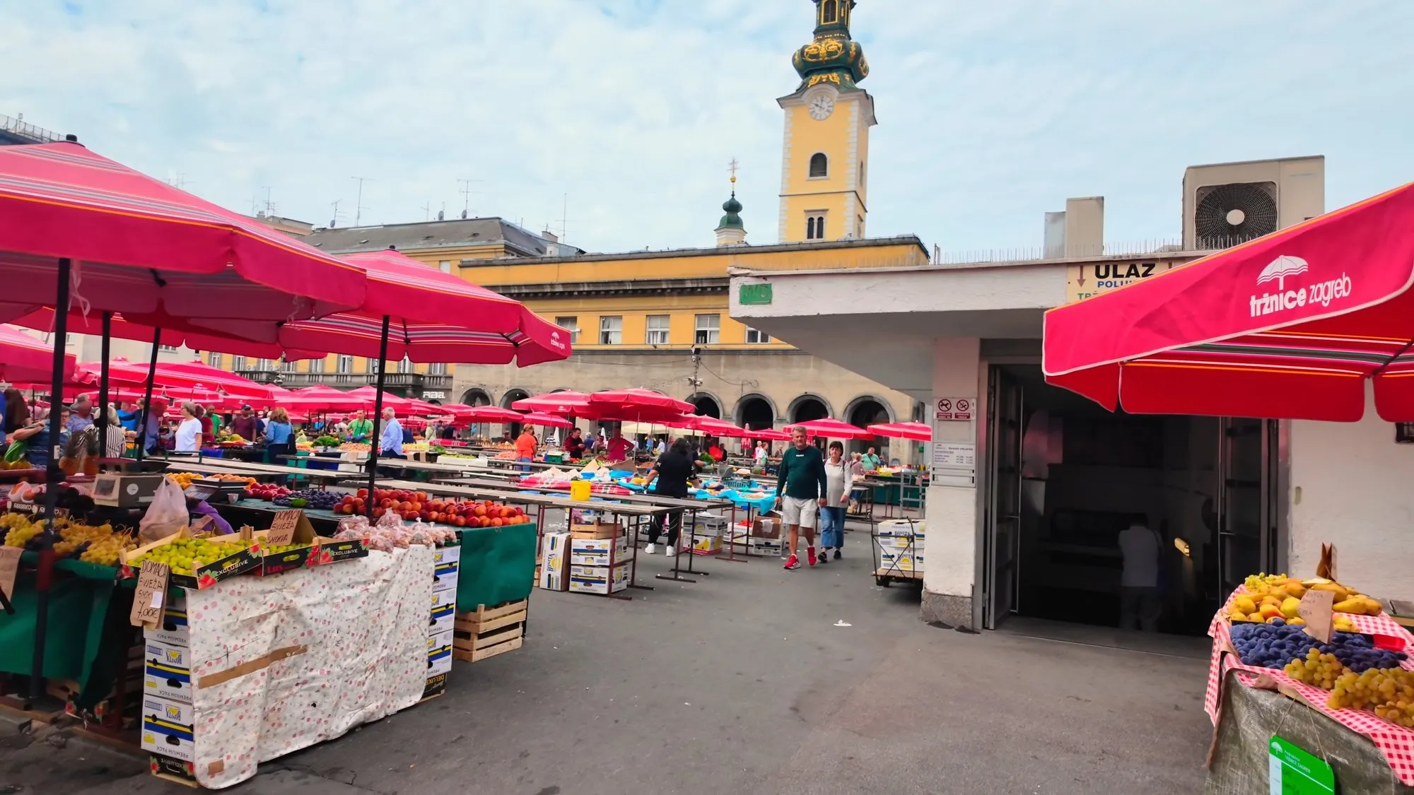 Red parasols over the open-air stalls of Dolac Market in Zagreb, with crates and tables of peaches, plums and grapes in the foreground and a yellow church tower behind