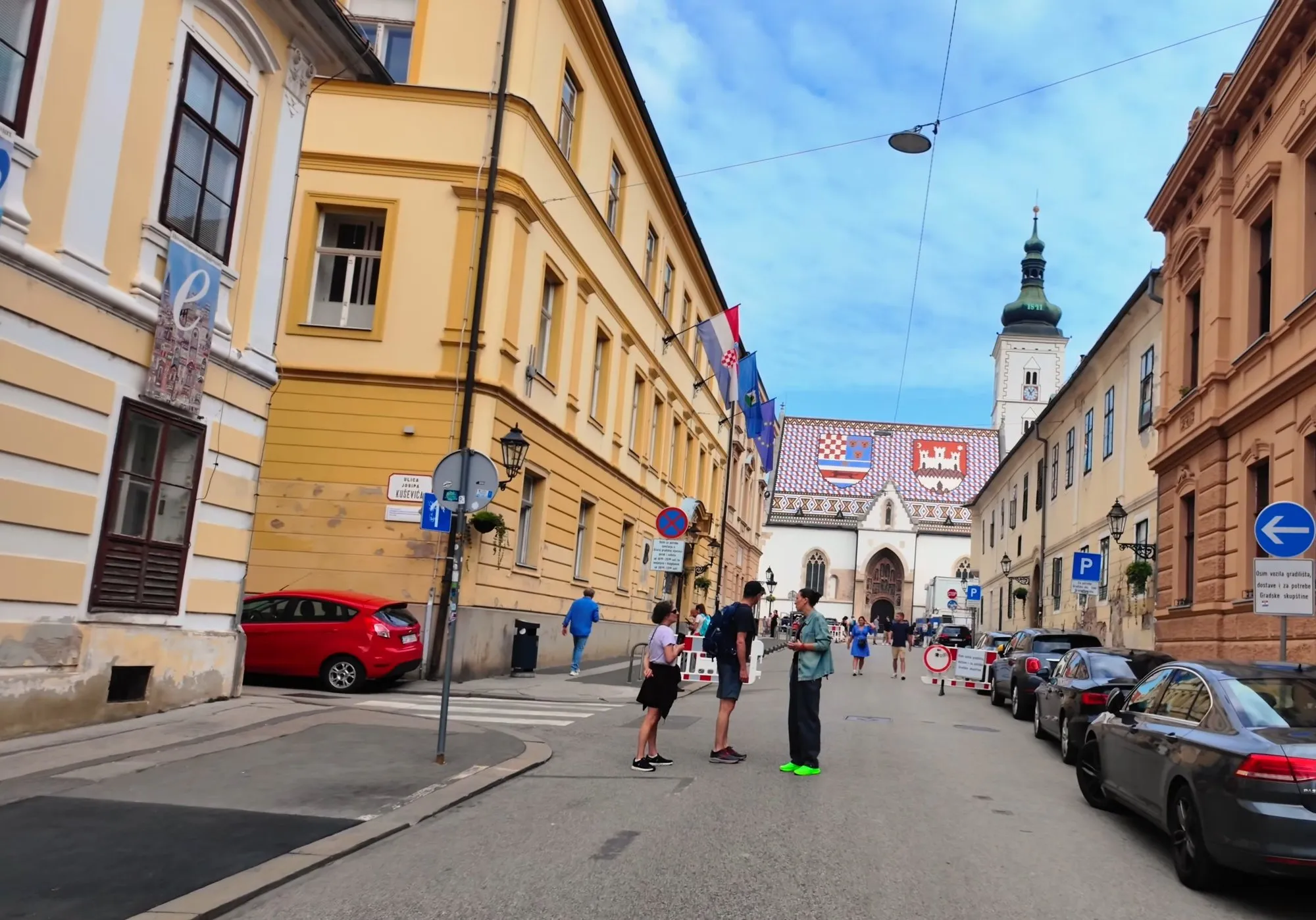 St Mark’s Church at the end of a street in Zagreb, with the Croatian-Dalmatian-Slavonian arms and the arms of the City of Zagreb in the tiles of its roof, and a few people on the street
