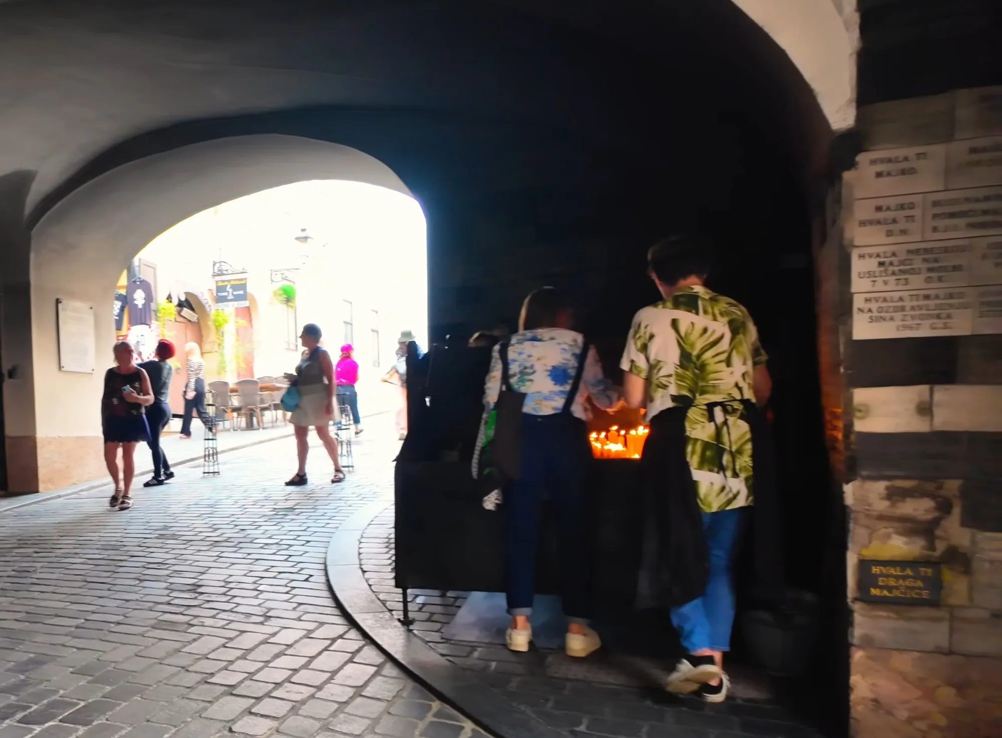 Candles burning at the shrine inside the Stone Gate in Zagreb, with two visitors seen from behind, a cobbled lane running out to a sunlit street and walls lined with small plaques reading HVALA, thank you