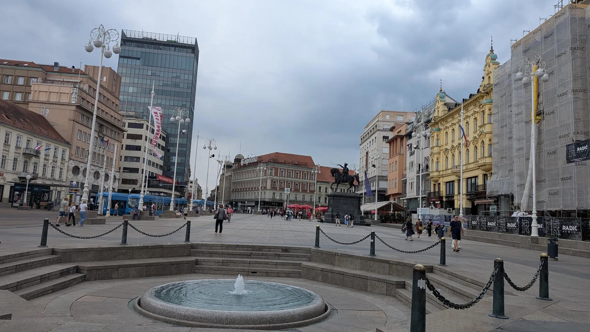 The low round basin of the Manduševac Fountain on Ban Jelačić Square in Zagreb, with the statue of Ban Jelačić and a scaffolded building behind it under a grey sky