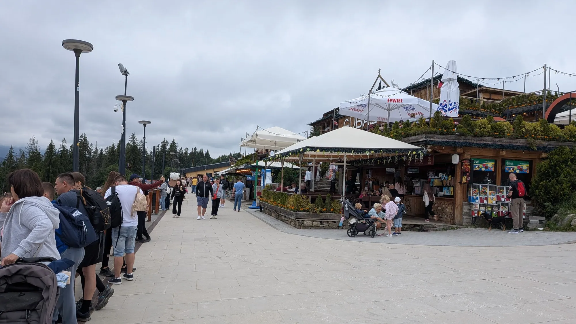 The wide paved promenade along the top of Gubałówka, with timber restaurants, café umbrellas and visitors walking under a cloudy sky